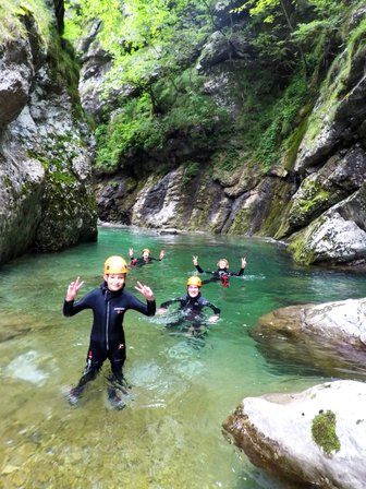 Eine Gruppe von Menschen in Neoprenanzügen genießt das Wasser eines klaren Flusses, umgeben von hohen Felsen und Bäumen.