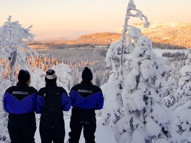Drei Personen in winterlicher Kleidung stehen auf einem schneebedeckten Hügel und blicken auf eine verschneite Landschaft.