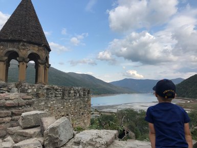 Ein Junge sitzt auf einer Mauer und blickt auf eine malerische Landschaft mit Bergen und einem ruhigen See.