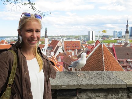 Eine lächelnde Frau steht an einer Mauer mit Blick auf die Altstadt von Tallinn und eine Möwe neben ihr.