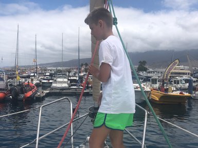 Ein Junge steht barfuß auf einem Segelboot und hält an einem Seil, während er den Hafen mit vielen Booten im Hintergrund betrachtet.