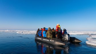 Eine Gruppe von Menschen in bunten Jacken steht auf einem Schlauchboot, umgeben von Eis und klarem blauen Himmel.