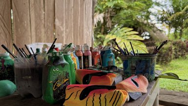 Bunte Malutensilien und Masken liegen auf einem Tisch, umgeben von üppiger tropischer Vegetation.