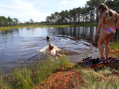 Drei Kinder genießen einen Sommertag am See, während zwei von ihnen im Wasser schwimmen und das dritte am Ufer steht.