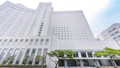 Ein modernes Hotelgebäude mit einer hellen Fassade und großen Fenstern, umgeben von gepflegten Bäumen und einem Parkplatz.