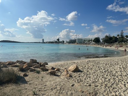 Ein sonniger Strand mit feinem Sand, klarem Wasser und Menschen, die sich am Ufer entspannen und schwimmen.