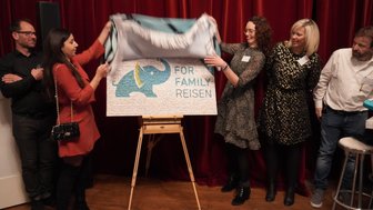 Eine Gruppe von Menschen enthüllt ein buntes Plakat mit einem Elefanten und dem Text 'FOR FAMILY REISEN' auf einer Leinwand.