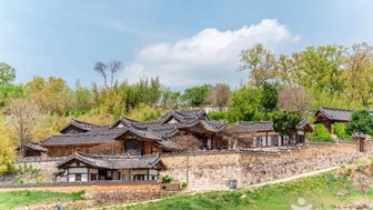 Traditionelle koreanische Häuser mit Ziegeldächern stehen in einer malerischen Landschaft, umgeben von Bäumen und sanften Hügeln.