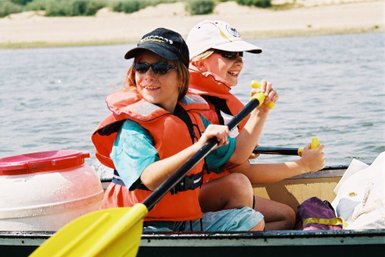 Zwei fröhliche Kinder in Schwimmwesten paddeln in einem Kanu auf einem ruhigen Gewässer, umgeben von einer malerischen Landschaft.