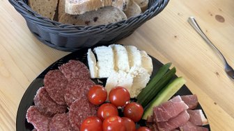Ein runder Teller mit verschiedenen Käsesorten, Wurst, Kirschtomaten und Gurken, umgeben von einem Korb mit Brotscheiben.