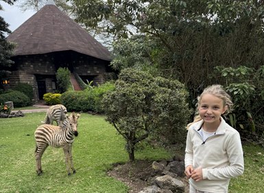 Ein Mädchen steht lächelnd in einem Garten, während ein Zebra in der Nähe grast, umgeben von üppigem Grün.