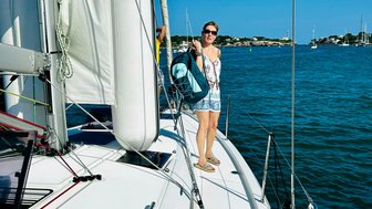 Eine Frau steht auf dem Deck eines Segelbootes, während sie eine Tasche trägt und den klaren blauen Himmel betrachtet.