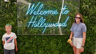 Ein Junge mit blauen Sonnenbrillen und ein Mädchen stehen vor einem leuchtenden 'Welcome to Hollywood'-Schild, umgeben von Pflanzen.