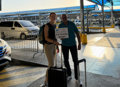 Eine Frau und ein Mann stehen am Flughafen, beide lächeln vor einem Schild mit der Aufschrift 'Für Familienreisen'.