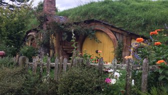 Ein charmantes Hobbit-Haus mit einer gelben Tür, umgeben von bunten Blumen und einem niedrigen Holzzaun.