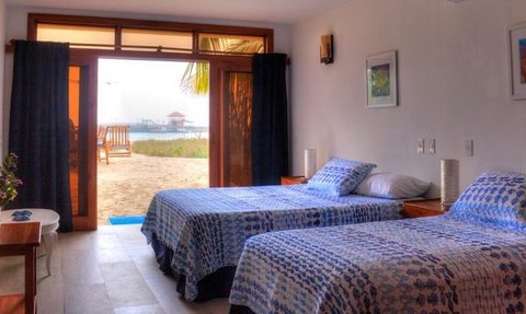 Ein helles Zimmer mit zwei Einzelbetten, blauen Bettdecken und Blick auf einen Strand durch große Fenster.