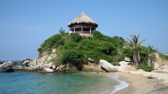 Ein malerisches Strandhaus mit Strohdach thront auf einer bewachsenen Klippe, umgeben von klarem Wasser und sanften Wellen.