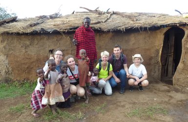 Eine Gruppe von Touristen und Maasai-Kindern posiert fröhlich vor einer traditionellen Lehmhütte in der afrikanischen Landschaft.