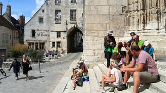 Eine Gruppe von Menschen sitzt auf Stufen, während andere Passanten in einer malerischen Stadt vorbeigehen.
