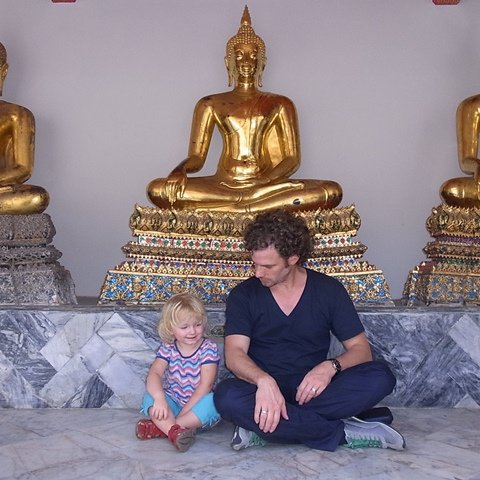 Ein Vater sitzt auf dem Boden und spielt mit seiner kleinen Tochter vor goldenen Buddha-Statuen in einem Tempel.