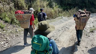 Drei Wanderer tragen große Körbe auf dem Rücken und gehen einen schmalen, steinigen Weg entlang, umgeben von üppiger Vegetation.