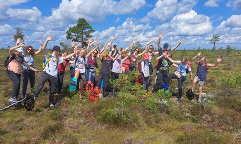 Eine Gruppe von Menschen mit Schneeschuhen posiert fröhlich in einer Moorlandschaft unter einem blauen Himmel mit Wolken.