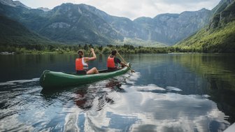 Zwei Personen paddeln in einem grünen Kanu auf einem ruhigen See, umgeben von majestätischen Bergen und üppigem Grün.