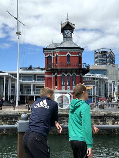 Zwei Jungen stehen am Wasser und schauen auf ein historisches Gebäude mit einer Uhr und einem roten Anstrich.