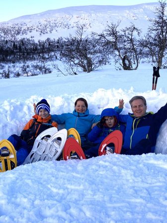 Eine Familie sitzt im Schnee, umgeben von schneebedeckten Bergen, und winkt fröhlich in die Kamera.