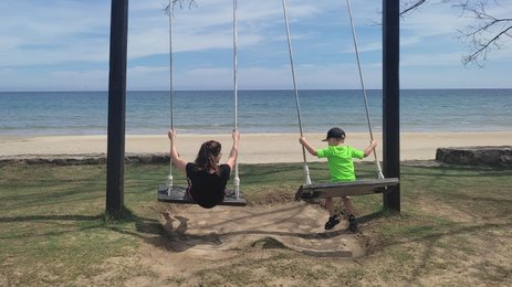 Eine Frau und ein Kind schwingen glücklich an einer Schaukel am Strand, umgeben von sanften Wellen und klarem Himmel.