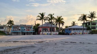 Ein ruhiger Strand mit sanften Wellen, umgeben von Palmen und bunten Häusern im Hintergrund, unter einem klaren Himmel.