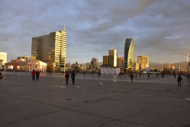 Die Stadt Ulaanbaatar wird von modernen Gebäuden und einem großen Platz mit Menschen belebt, während die Sonne untergeht.