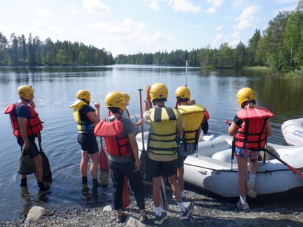 Eine Gruppe von sechs Personen in Schwimmwesten und Helmen steht am Ufer eines ruhigen Gewässers, bereit zum Rafting.