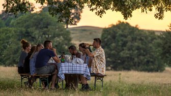 Eine Gruppe von Freunden sitzt an einem Tisch im Freien, umgeben von Natur, und genießt ein gemeinsames Essen bei Sonnenuntergang.