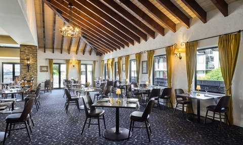 Ein elegantes Restaurant mit Holzbalkendecke, stilvollen Tischen und Stühlen, umgeben von großen Fenstern und goldenen Vorhängen.