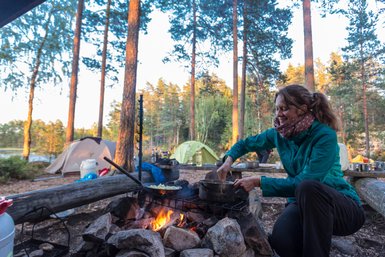 Eine Frau kocht am Lagerfeuer in einem Waldcamp, umgeben von Zelten und Bäumen, während die Sonne untergeht.