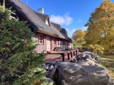 Ein traditionelles Reetdachhaus mit einer Holzveranda, umgeben von buntem Herbstlaub und einem blauen Himmel.