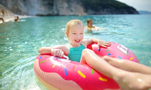 Ein fröhliches Mädchen mit blonden Locken genießt das Wasser in einem bunten Donut-Floatie an einem sonnigen Strand.