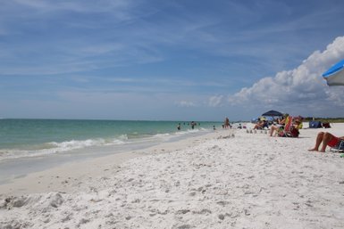 Ein sonniger Strand mit feinem, weißem Sand, wo Menschen im Wasser spielen und sich entspannen.