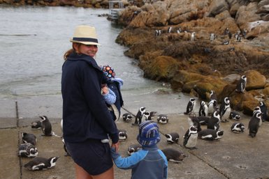 Eine Frau mit einem Hut hält ein Baby, während ein Junge an ihrer Hand steht und Pinguine im Hintergrund beobachtet.