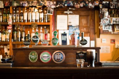 Eine Holzbar mit verschiedenen Bierzapfhähnen, darunter Guinness und Heineken, vor einer Wand voller Flaschen und Erinnerungsfotos.