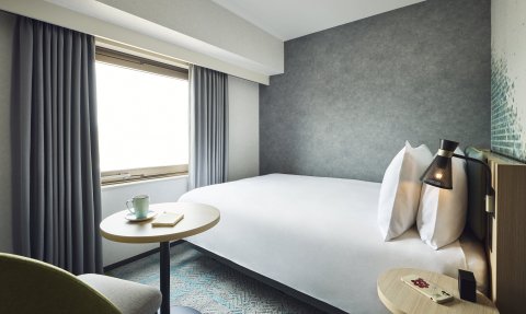 Ein modernes Hotelzimmer mit einem großen Bett, einem kleinen Tisch und einem Fenster, das viel Tageslicht hereinlässt.