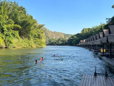 Kinder schwimmen an Floathouses am Flussufer in ruhiger Wasserlandschaft vorbei - Thailand mit Kindern