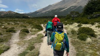 Drei Wanderer gehen auf einem schmalen Pfad durch die grüne Landschaft, umgeben von Bergen und einem klaren Himmel.