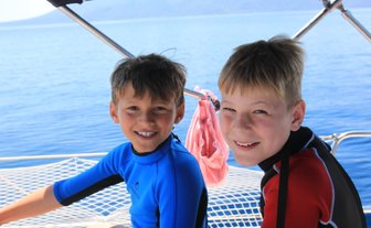 Zwei fröhliche Jungen in Neoprenanzügen sitzen auf einem Boot und genießen die Sonne und das Wasser um sie herum.