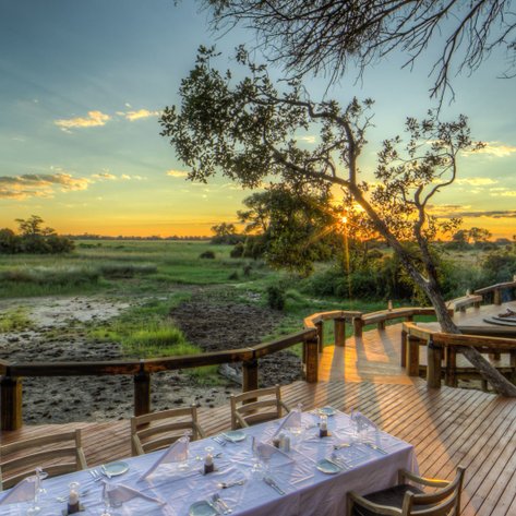 Ein eleganter Tisch mit weißer Tischdecke steht auf einer Holzterrasse, umgeben von der malerischen Landschaft Botswanas bei Sonnenuntergang.