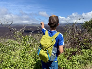 Ein Junge mit einem grünen Rucksack steht auf einem Hügel und zeigt auf die weite Vulkanlandschaft vor ihm.