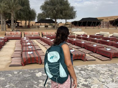 Eine Person mit einem Rucksack steht auf einem gepflasterten Weg und blickt auf eine Wüstenlandschaft mit Zelten.