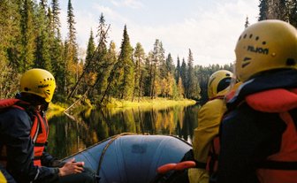 Eine Gruppe von Menschen in einem Schlauchboot, umgeben von hohen Bäumen und einem ruhigen Fluss in Finnland.