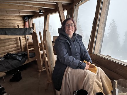 Eine lächelnde Frau sitzt in einer gemütlichen Holzhütte, umgeben von Fenstern mit Blick auf neblige Bäume.